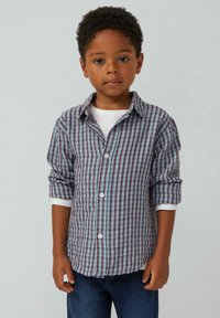 Gecurlerd shirt met een ruitjespatroon in blauw, bordeauxrood en wit; lange mouwen opgerold; gedragen over een wit t-shirt; donkere spijkerbroek.