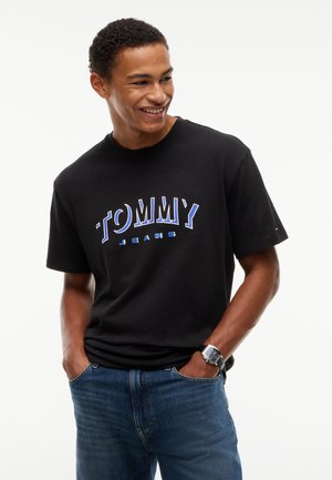 Jeune homme souriant portant un t-shirt noir Tommy Jeans, un jean bleu et une montre-bracelet, les mains dans les poches, sur fond blanc.