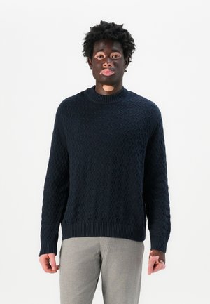 Navy blue knitted sweater featuring a textured wave pattern, ribbed neckline, and cuffs. Paired with light gray trousers.