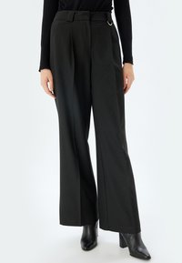 Wijdvallende zwarte broek met verticale streepjes, een taille met riem en een zilveren hardware ring, en een licht gestructureerde stof.
