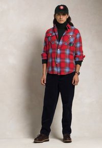 Röd rutmönstrad flanellskjorta med bröstfickor, svart polotröja, marinblå byxor och bruna skor. Grön keps med logodetalj. Modellen står upprätt.