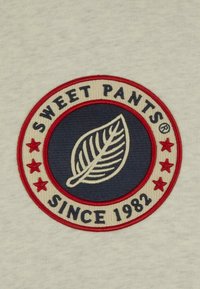 Patch brodé circulaire avec un fond bleu foncé, un motif de feuille et des étoiles rouges. Texte : "SWEET PANTS®" et "SINCE 1982" en crème.