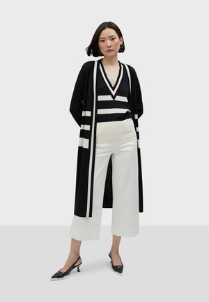 Un cárdigan largo a rayas en blanco y negro sobre una blusa de escote en V, combinado con pantalones anchos blancos y unos zapatos de tacón kitten con punta negra.