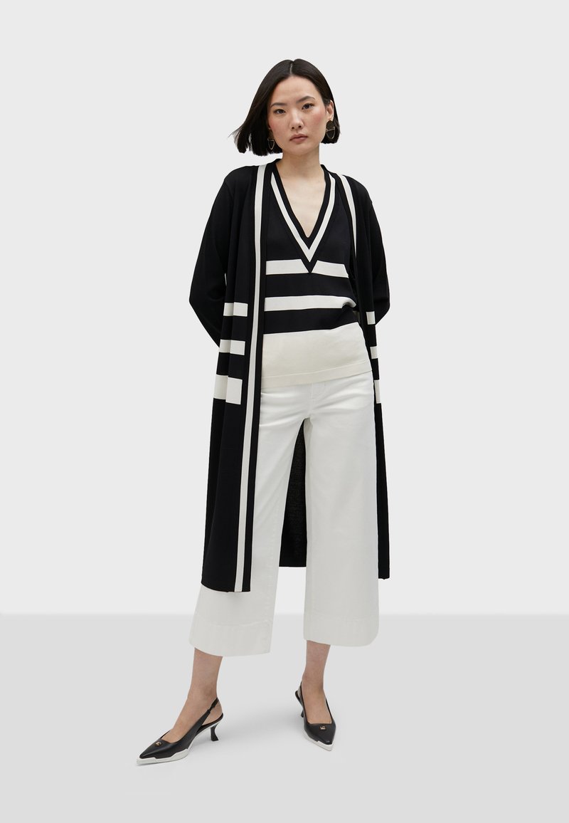 Svartvit randig lång cardigan över en svart v-ringad topp och vita vida byxor, kombinerat med svarta slingbackskor med klack.