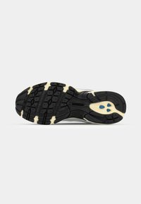 Semelle en caoutchouc noire et beige d'une chaussure de course New Balance avec des motifs de la bande de roulement et des points de traction bleus près du talon.