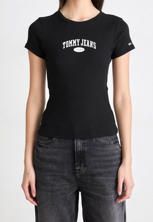 Czarny prążkowany T-shirt z krótkimi rękawami, z białym napisem "TOMMY JEANS" i logo na klatce piersiowej. Noszony z ciemnoszarymi jeansami.