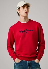 Joven vistiendo un sudadera roja de Pepe Jeans, jeans azules y una gorra blanca, mirando de lado con las manos en los bolsillos, fondo gris neutro.
