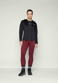 Crna zip-up sportska jakna s logotipom, siva oblikovana majica, bordo tajice s bočnim logotipom i crne cipele s bijelim detaljima.