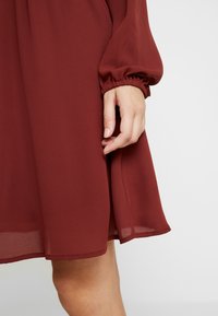 Vestido burdeos a la altura de la rodilla con mangas largas y fruncidas, hecho de un tejido ligero y transparente con una textura suave. Mano visible en el dobladillo.