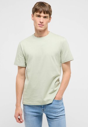 Junger Mann steht mit einer Hand in der Tasche, trägt ein hellgrünes kurzärmeliges T-Shirt und hellblaue Jeans vor einem schlichten Hintergrund.