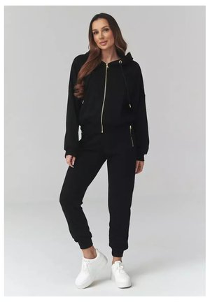 Kobieta stojąca w czarnej bluzie z kapturem na zamek, czarnych spodniach dresowych i białych trampkach na jednolitym szarym tle.