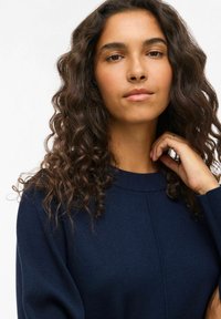 Pull en tricot bleu marine avec un col rond, des poignets côtelés et un détail de couture devant. Le modèle a de longs cheveux bouclés effleurant légèrement son cou.