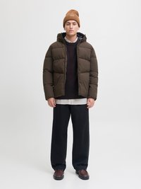 Rjava napihnjena jakna s kapuco, nošena čez temno pulover in belo rebrasto majico. Ujemajo se s črnimi širokimi hlačami in rjavimi čevlji.