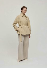 Beige trench coat med bälte i midjan, stor krage och knappdetaljer, i kombination med ljusbeiga utsvängda byxor och krämfärgade skor.