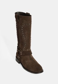 Botte en cuir suédé marron, montant jusqu'au genou, avec des clous décoratifs en argent, une semelle plate et une sangle à la cheville. Présente un embout légèrement arrondi et des coutures décoratives.