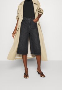 Musta denimin culottes-housut, korkeavyötäröiset ja leveälahkeiset. Yhdistetty mustaan ribbineulepaitaan ja beigeen trenssitakkiin. Mustat korolliset varvassandaalit.