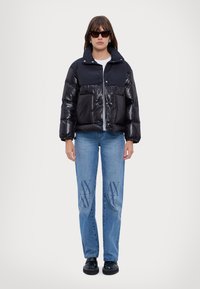 Giacca piumino nera e blu con zip, polsini elasticizzati e due tasche frontali indossata sopra una maglietta grigia e un paio di jeans blu con dettagli del logo.