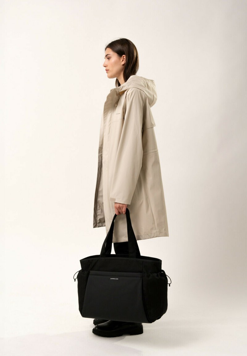 Impermeabile beige con cappuccio, lungo fino al ginocchio. Borsa a tracolla nera con due manici robusti e tasca frontale. Il modello si trova di profilo su uno sfondo semplice.