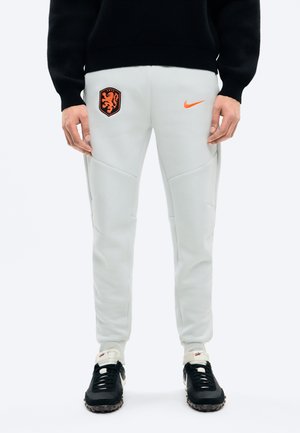 Białe spodnie sportowe z pomarańczowym herbem lwa Holandii oraz pomarańczowym logo Nike, noszone z czarnym swetrem i czarno-białymi butami Nike.