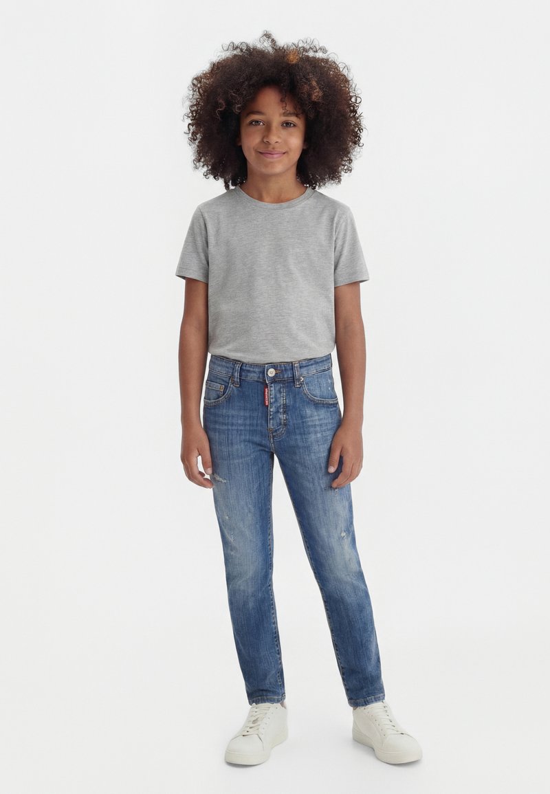 Enfant aux cheveux bouclés debout, portant un t-shirt gris à manches courtes, un jean bleu et des baskets blanches sur un fond blanc uni.