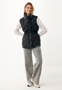 Černá prošívaná vesta s vysokým límcem, páskem v oblasti pasu a lesklou texturou, doplněná o světle šedé široké džíny a tenisky. Jednoduché pozadí.