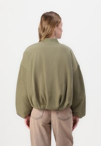 Olijfgroene, oversized jas met een geribbelde kraag, verlaagde schouders en een gathered zoom, gecombineerd met lichtbruine, hoog-taille broeken.