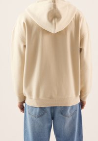 Beige hoodie met een capuchon, gedragen door een persoon die van zich af kijkt, gecombineerd met lichtblauwe denim jeans tegen een effen achtergrond.