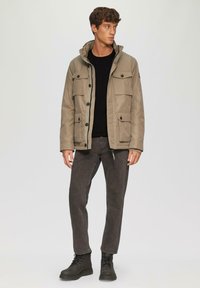 Beige jas met dubbele borstzakken, zwarte knopen en verstelbare capuchon. Draagt over een zwarte sweater en gecombineerd met donkergrijze jeans en zwarte laarzen.