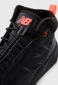 Sneaker New Balance noir avec des détails en mesh et en cuir, logo rouge et système de laçage ajustable, et tirant arrière rouge, présenté en gros plan.