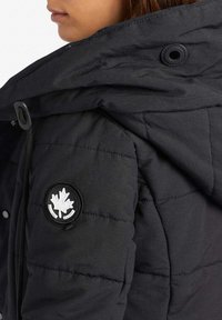 Svart pufferjacka med huva, knappstängning och en cirkulär svart och silver logotyp med ett lönnblad på ärmen.