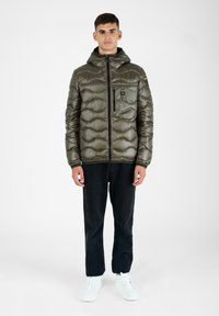 Olivgrüne Puffermantel mit Kapuze, gestepptem Design, Reißverschluss-Vordertasche und schwarzen Akzenten, kombiniert mit schwarzen Hosen und weißen Turnschuhen.