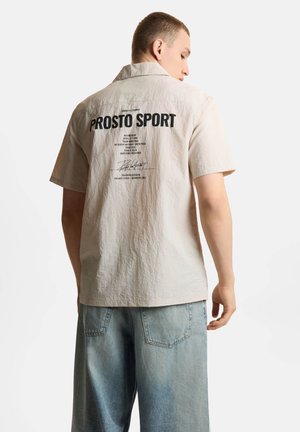 Młody mężczyzna zwrócony tyłem, ubrany w beżową koszulę z krótkim rękawem z napisem „PROSTO SPORT” oraz jasnoniebieskie dżinsy.