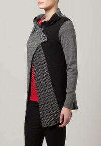 Asymmetrisk cardigan med en blandning av svarta och grå tyger, texturerade stickade mönster, hög krage och knappstängning fram.