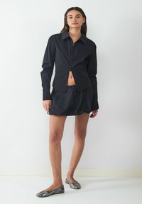 Schwarze Longsleeve-Button-up-Bluse mit verkürztem Saum, kombiniert mit einem schwarzen, gefalteten Minirock und glänzenden grauen Ballerinas. Glatte Stofftexturen.