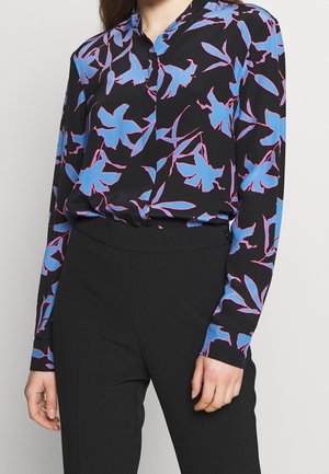 Femme portant un chemisier noir à boutons avec un imprimé floral bleu et rose, associé à un pantalon noir taille haute, debout les mains détendues.