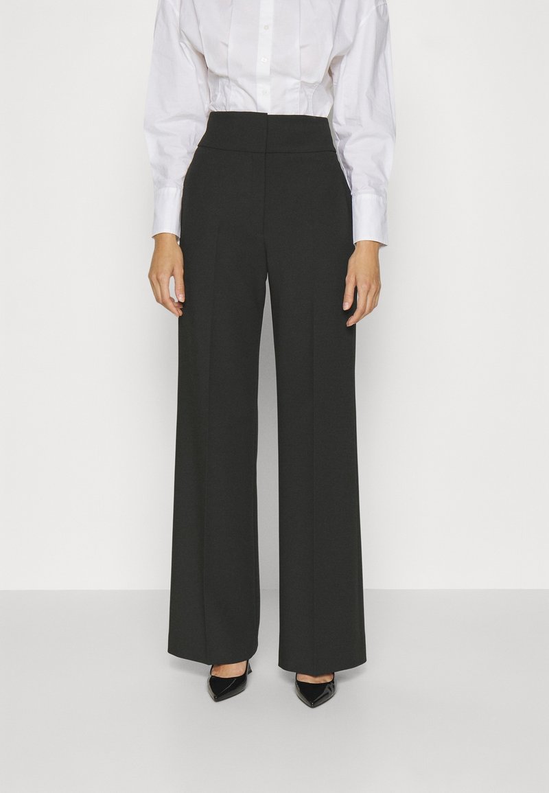 Femme portant un pantalon noir taille haute à jambes larges, une chemise blanche boutonnée et des talons noirs pointus, debout devant un fond clair uni.
