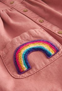 Chemise en tissu rose avec détails de boutons, ornée d'un patch arc-en-ciel multicolore brodé sur la poche poitrine gauche.