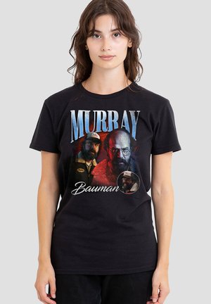 Woman wearing black T-shirt featuring three images of a man with glasses and beard, with text "Murray Bauman" on the front.