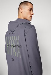 Grå hettegenser med en tekstdesign i svart og grønt på baksiden. Laget av glatt stoff med en avslappet passform.