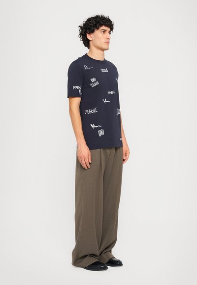 Marineblå bomulds t-shirt med hvid grafisk tekst og mønstre; parret med brede, brune bukser og sorte sko. Modellen står sidelæns.