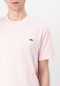 Kurzärmliges, hellrosa Baumwoll-T-Shirt mit grünem gesticktem Krokodil-Logo auf der linken Brust. Klassischer Rundhalsausschnitt.
