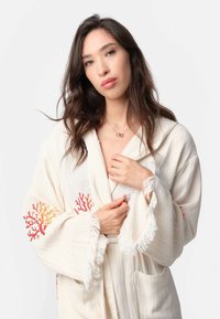 Ľahký krémový župan s strapcami, s koralovými výšivkami v červenej a oranžovej farbe, dvoma prednými vreckami a kapucňou.