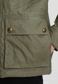 Olivově zelená bunda s hladkým povrchem, velkou přední kapsou se zapínáním na knoflík a kontrastním prošíváním.