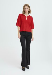 Crvena pletena majica s dekolteom u obliku suza, u kombinaciji s crnim širokim hlačama i crnim cipelama s šiljastim nosom. Majica ima kratke rukave i ležeran kroj.