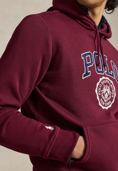 Άνδρας που φοράει μπουφάν Polo Ralph Lauren μπορντό με λογότυπο στο στήθος και μικρό λευκό άλογο στο μανσέτα του μανικιού, με το χέρι στην τσέπη.