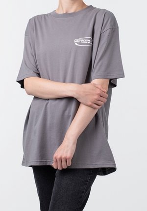 Osoba nosząca luźny szary t-shirt z białym tekstem na klatce piersiowej, stojąca ze skrzyżowanymi ramionami, w połączeniu z czarnymi spodniami na jednolitym tle.