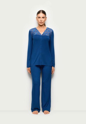 Frau steht barfuß und trägt ein blaues, langärmliges, geknöpftes Oberteil mit Spitzen-Details und passende weite Hosen vor einem einfarbigen Hintergrund.