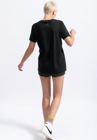 Zwarte katoenen t-shirt met een ronde hals en korte mouwen; heeft een kleine schaarafbeelding op de bovenrug. Gepaard met olijfgroene shorts.