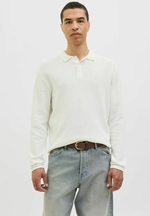 White long-sleeve polo shirt with a textured knit fabric, a soft collar, and a two-button placket, paired with light blue denim trousers.