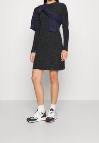 Abito grigio a maniche lunghe con una vestibilità rilassata, abbinato a sneakers multicolori e calze bianche. Un maglione blu navy è annodato sulle spalle.